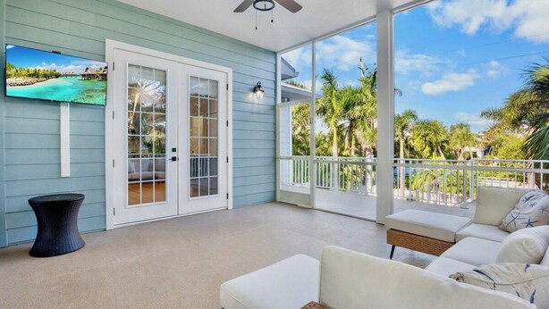 A covered porch with a white sofa, a television mounted on the wall, and a view of palm trees. - New! Gorgeous home on AMI. Heated pool! (Anna Maria)