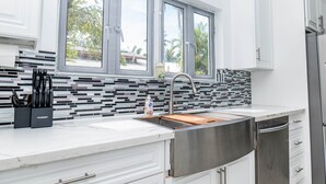 A modern kitchen with white cabinetry, a stainless steel sink, and a window with a view of palm trees. - Beachfront Luxury in Cable Beach West - Sleeps 6+ (Nassau)