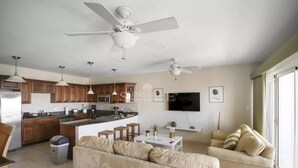 Una cocina moderna con armarios de madera, una isla central e iluminación colgante. Hay una zona de estar con un sofá color beige y un televisor montado en la pared.