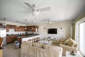A modern kitchen with wooden cabinets, a central island, and pendant lighting. There is a living area with a beige sofa and a TV mounted on the wall. - Caribbean Getaway: 2BR Condo at Rainbow Beach Club (Cupecoy)