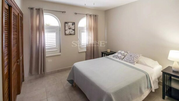 Un dormitorio con cama, dos ventanas con cortinas, un ventilador de techo y una mesita de noche con lámpara.