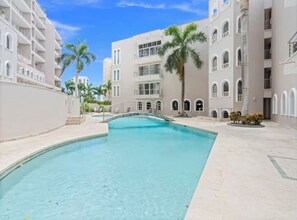 A swimming pool area with a clear blue pool, surrounded by multi-story buildings with balconies and arched windows. - Caribbean Getaway: 2BR Condo at Rainbow Beach Club (Cupecoy)