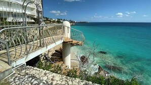 Un hotel costero con balcón que da a aguas turquesas, una escalera que conduce a los pisos superiores y una vista despejada del mar.