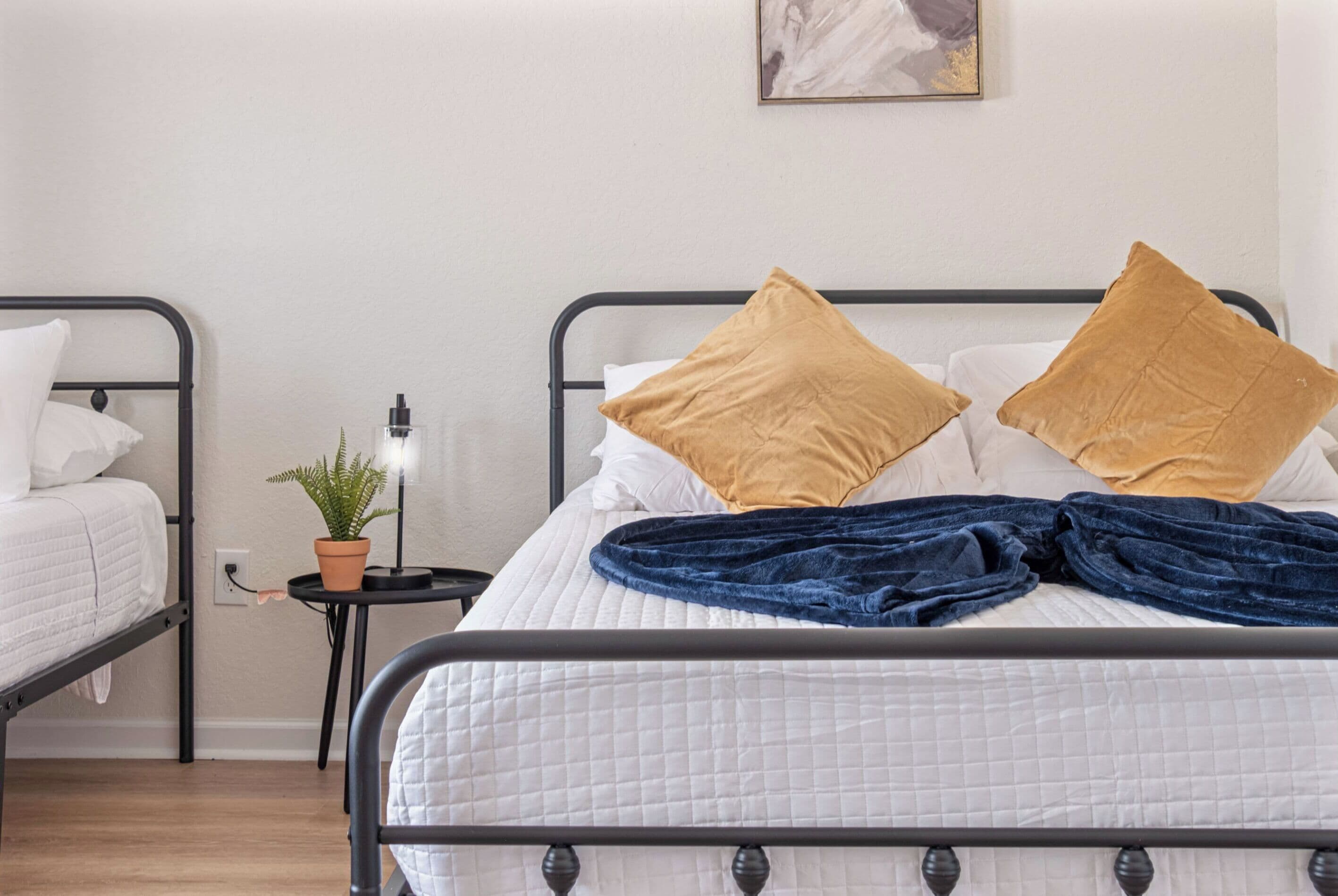 Ein Doppelbett mit weißer gesteppter Bettdecke und zwei Kissen, ein kleiner Nachttisch mit Lampe und Topfpflanze sowie ein Wandbild über dem Bett.