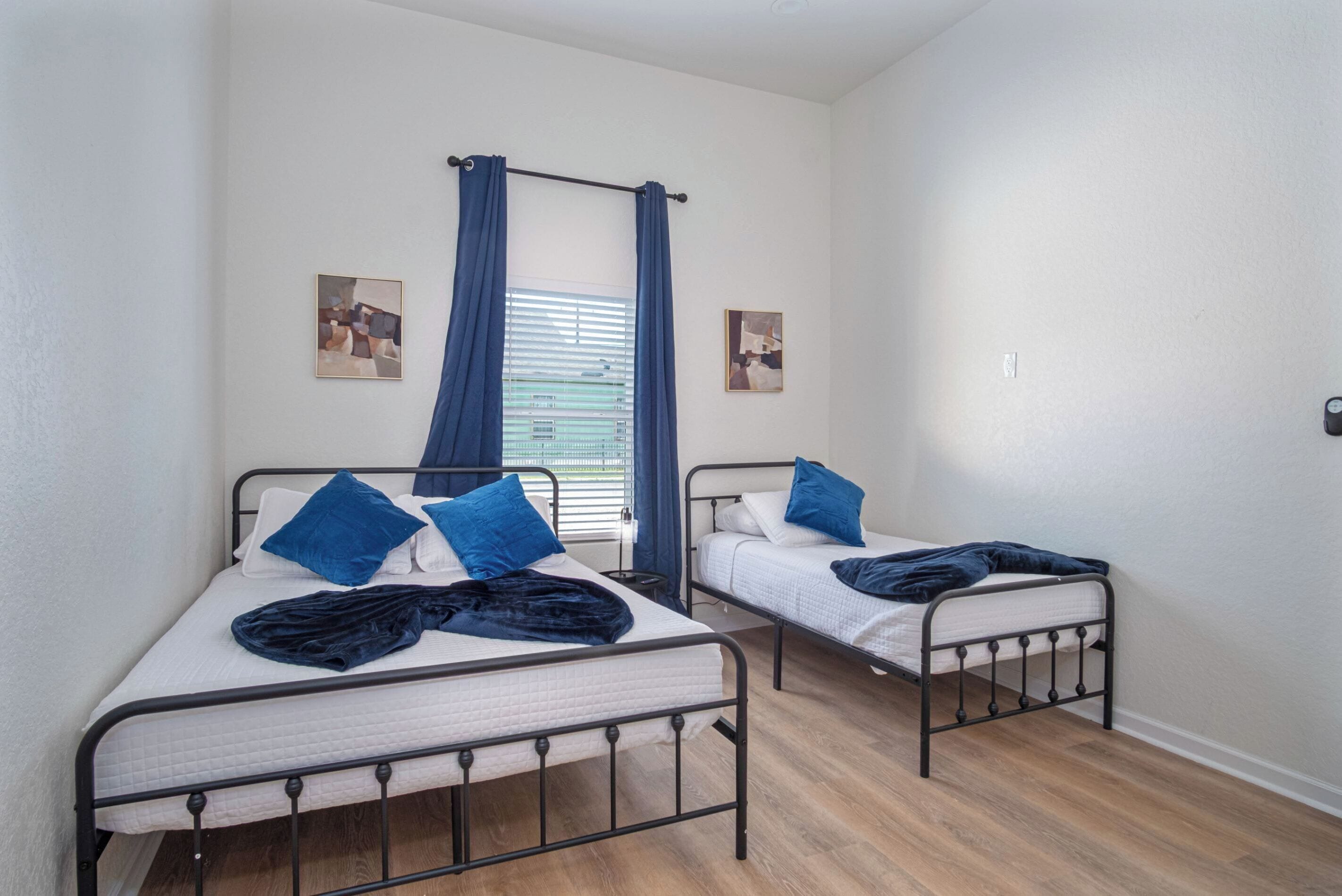 Een kamer met twee eenpersoonsbedden, elk met een blauwe deken en kussen, een raam met blauwe gordijnen en twee foto's in lijsten aan de muur.