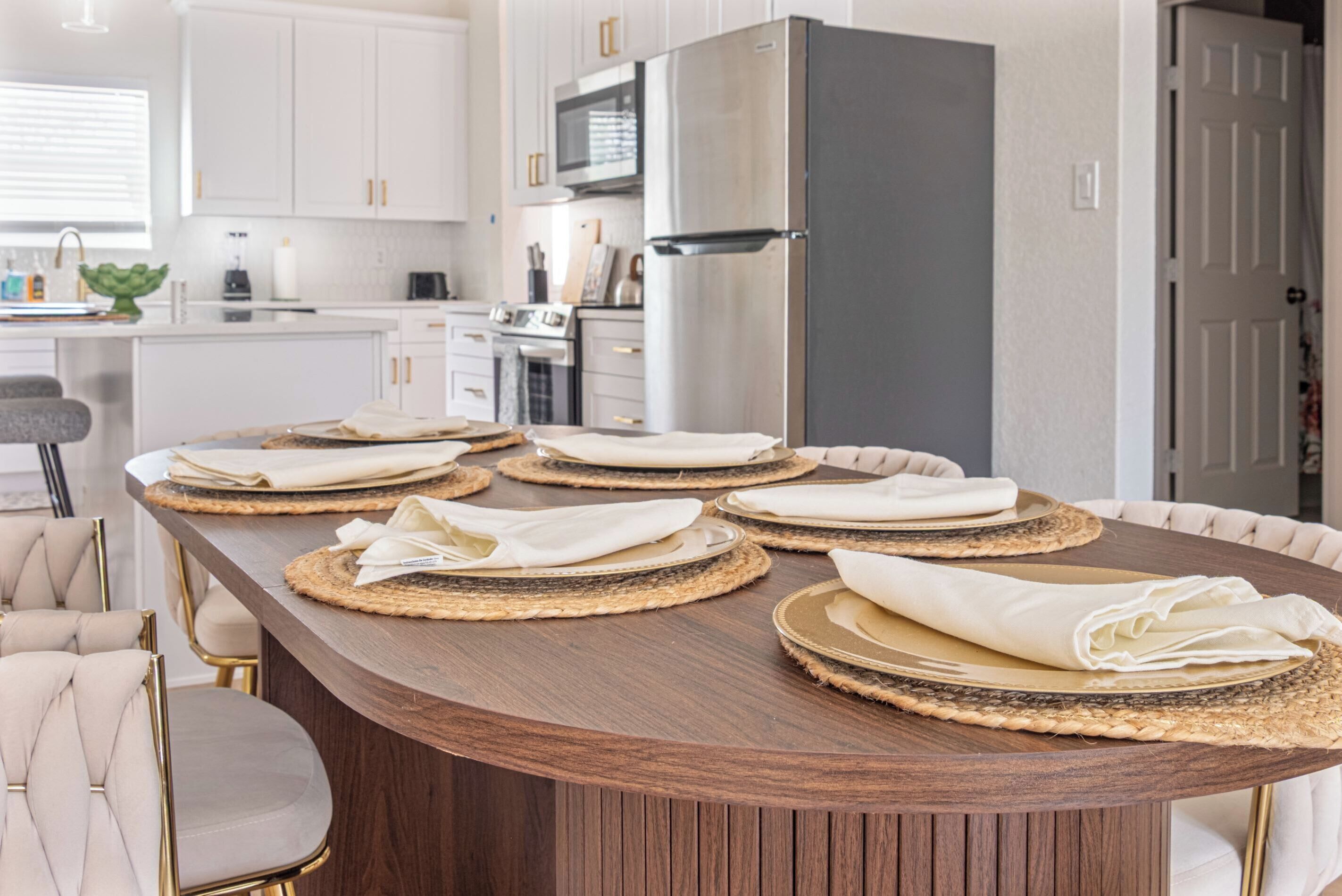A modern kitchen with white cabinets, a stainless steel refrigerator, and a dining table set with placemats and folded napkins.