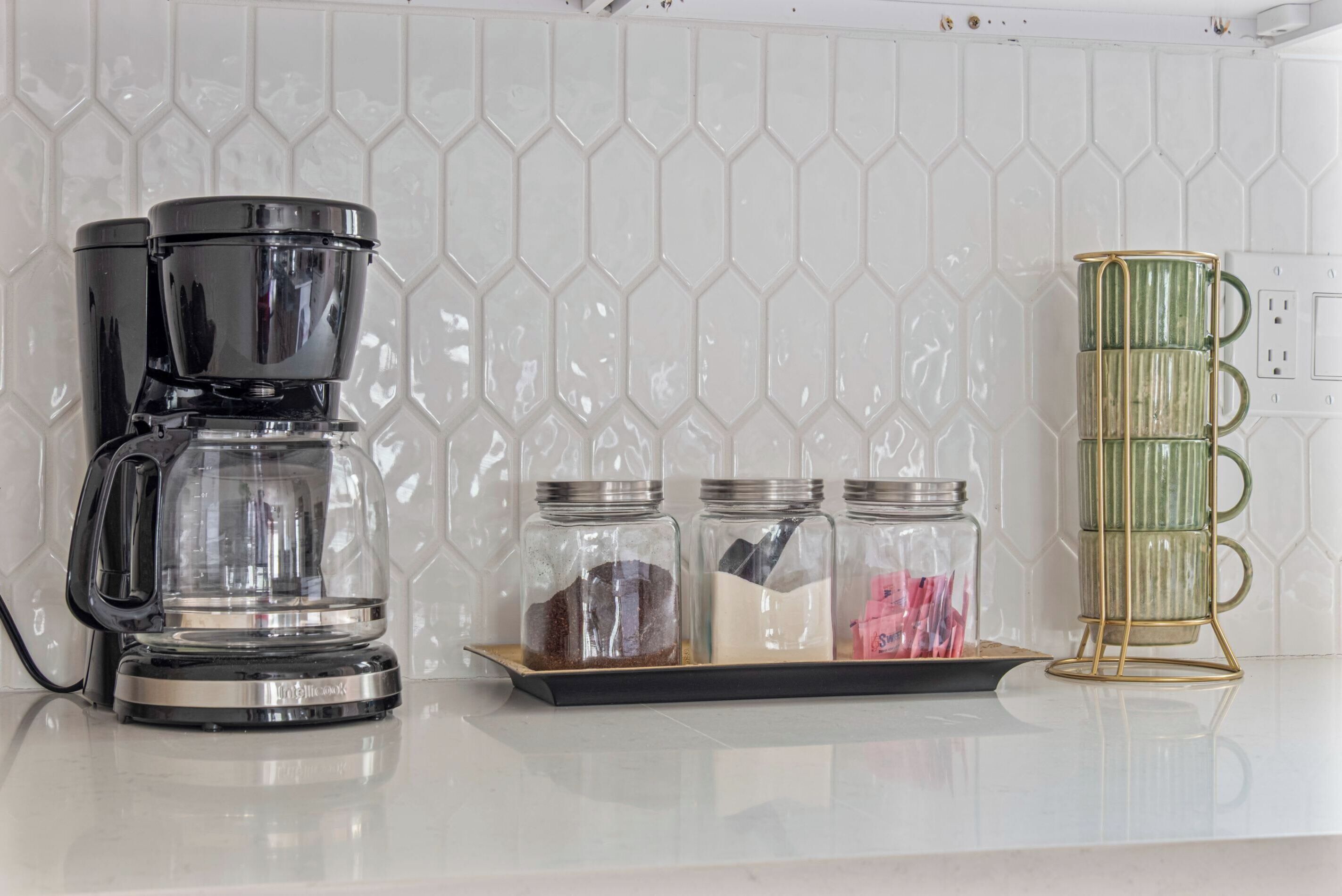 Una cocina con cafetera, tres frascos de vidrio sobre una bandeja y una taza verde en un pedestal.