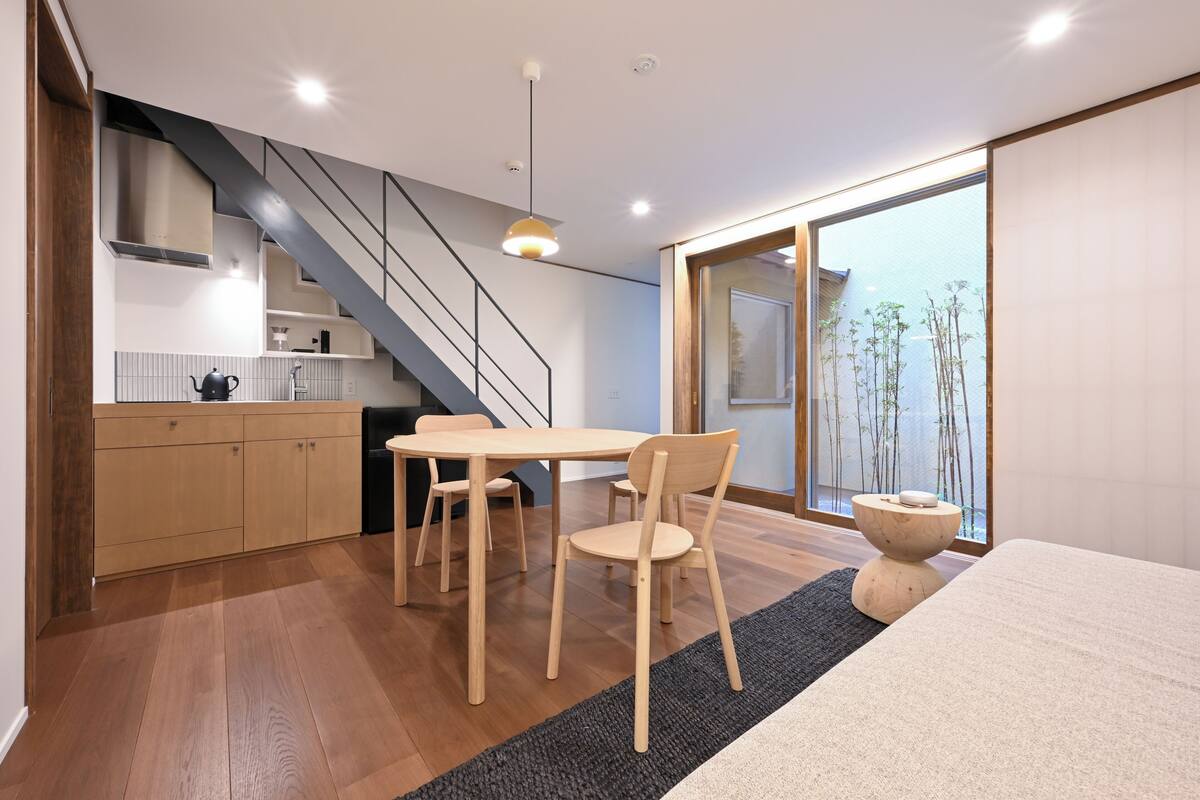 A modern kitchen with wooden cabinets, a dining table with chairs, a staircase, and a large window with a view of bamboo outside.