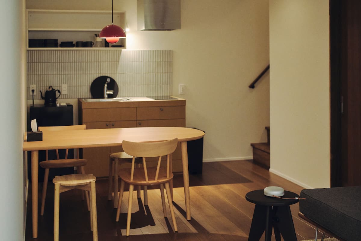 A compact kitchen with a wooden table, two chairs, a small stool, and a black side table with a white object.