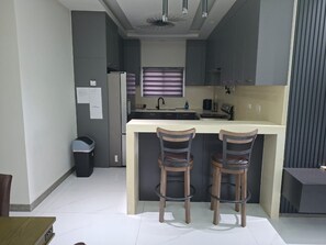 Eine moderne Küche mit einer zentralen Kochinsel, zwei Barhocker, einem Kühlschrank und einem Fenster mit Jalousien.