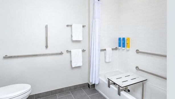Une salle de bain avec des toilettes blanches, une douche avec un rideau blanc, des barres d'appui et un sèche-mains.