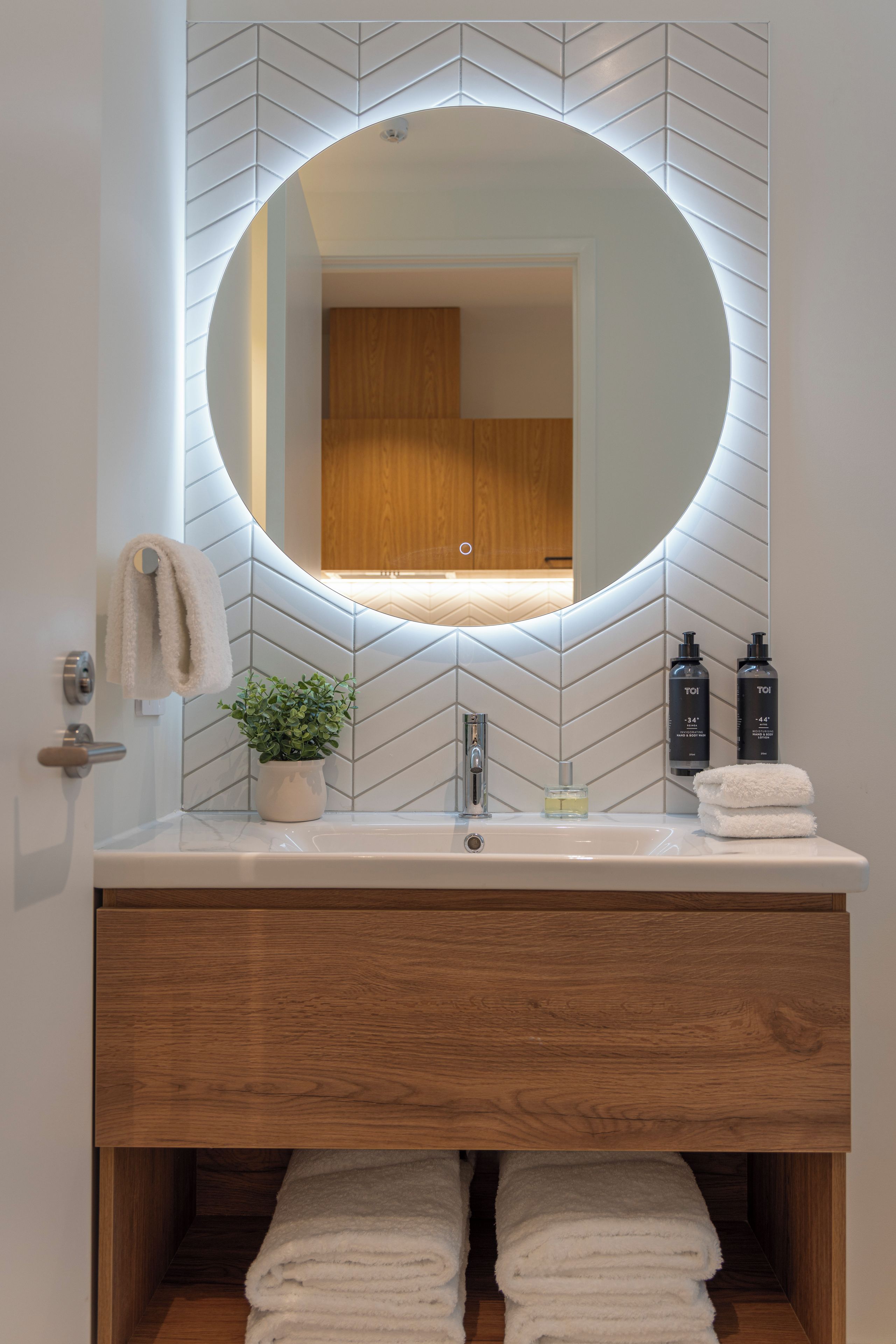Un bagno con uno specchio tondo, un mobiletto in legno, una pianta in vaso e articoli da toilette.