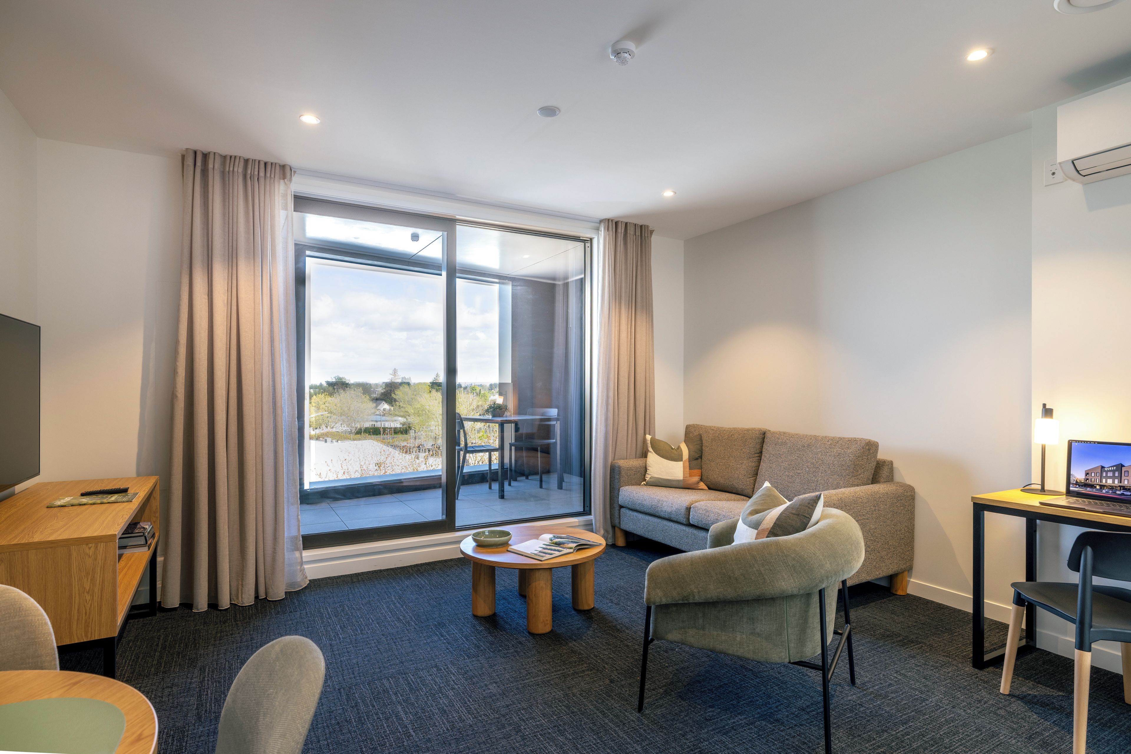 A modern hotel room with a sofa, a desk, a laptop, and a dining table.