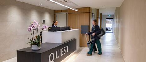 Un comptoir d’accueil portant l’inscription « QUEST », agrémenté d’une orchidée en pot, une réceptionniste, et un couloir avec une enseigne « BOUTIQUE ».