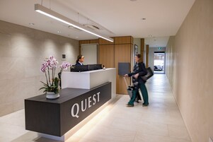 Una recepción con el rótulo 'QUEST', una orquídea en maceta, una recepcionista y un pasillo con el letrero 'BOUTIQUE'.