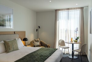Habitación de hotel con una cama, dos sillas, una mesita con una planta y bebidas, y una ventana con cortinas.