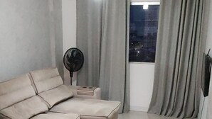 A modern living room with a sectional sofa, a standing fan, and a view of the city at night through large windows. - Comfortable, fully equipped 3-bedroom apartment with garage and 24-hour security. (Belém)
