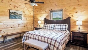 A wooden cabin bedroom with a bed, bench, two bedside tables, a ceiling fan, and a window. - Bear Monkey Lodge - Luxury 4 Bedroom with Hot Tub + Fireplaces + Scenic Views! (Sevierville)
