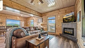 A cozy log cabin interior with a leather sofa, a fireplace, and large windows offering scenic views. - Bear Monkey Lodge - Luxury 4 Bedroom with Hot Tub + Fireplaces + Scenic Views! (Sevierville)