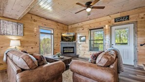 A cozy cabin interior with wooden walls, a fireplace, two leather sofas, and a ceiling fan. - Bear Monkey Lodge - Luxury 4 Bedroom with Hot Tub + Fireplaces + Scenic Views! (Sevierville)