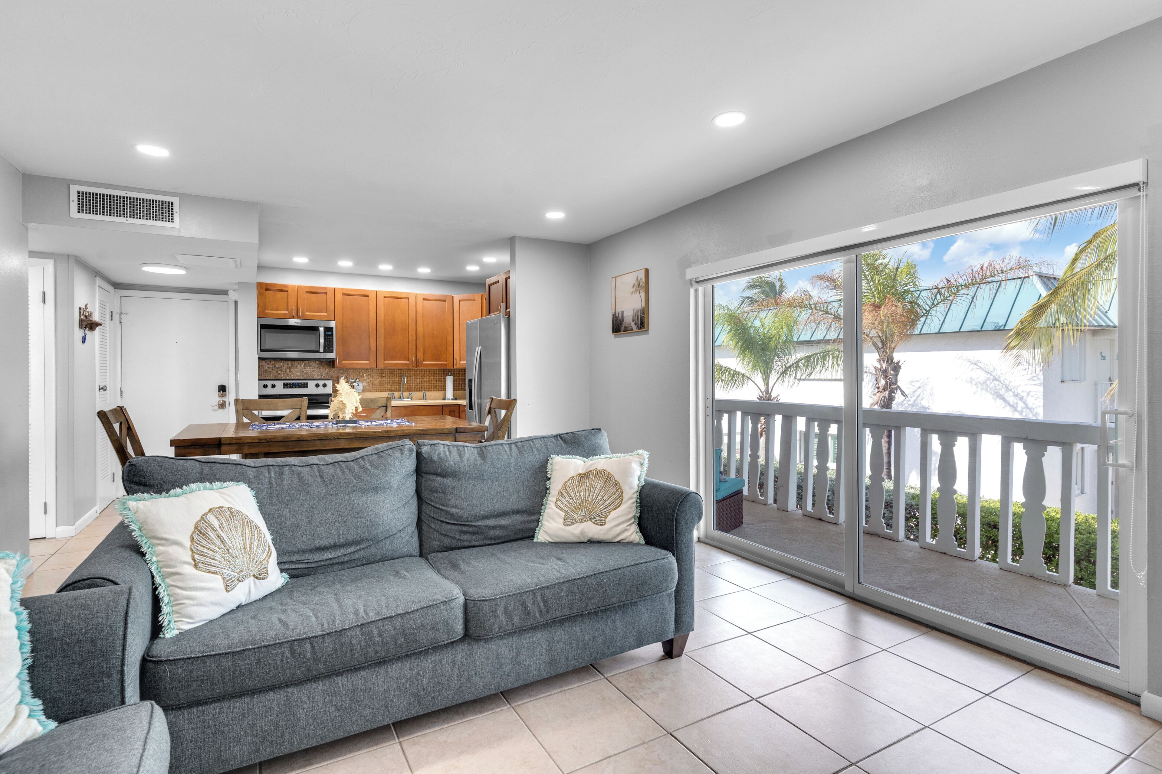 A modern living room with a grey sofa, a dining area with wooden cabinets, and a large sliding glass door leading to a balcony with a view of palm trees.