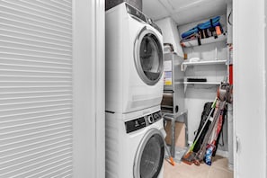A compact laundry room with a front-loading washer and dryer, a small shelf with cleaning supplies, and a vacuum cleaner. - Sombrero Beach Getaway - Pool (Marathon)
