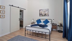 A bedroom with a bed, bedside tables, a nightstand, a painting, and a door leading to a bathroom. - Bright and Clean 3 Bedroom Near Fun Riverwalk (San Antonio)