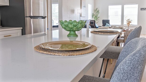 A modern kitchen with a stainless steel refrigerator, a central island with a decorative green vase, and a dining area with grey upholstered chairs. - Bright and Clean 3 Bedroom Near Fun Riverwalk (San Antonio)