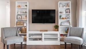 Una sala de estar con un televisor de pantalla plana, dos sillones grises y estanterías blancas decoradas con objetos ornamentales.