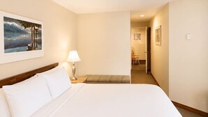 Ett hotellrum med en stor säng, en sänglampa, en tavla på väggen och en hall som leder till ett annat rum.