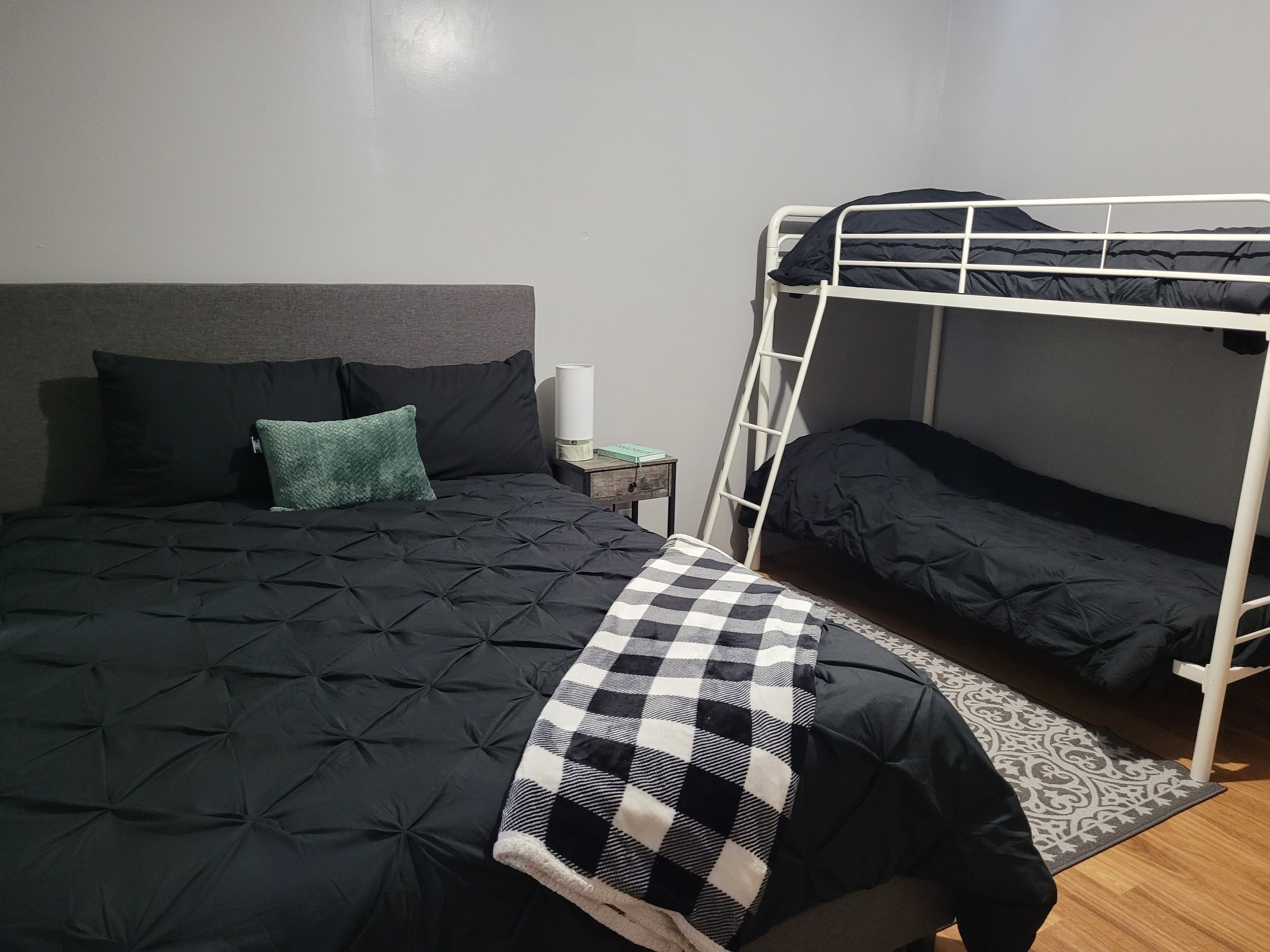 A bunk bed with a checkered blanket, a bedside table with a lamp, and a grey headboard.