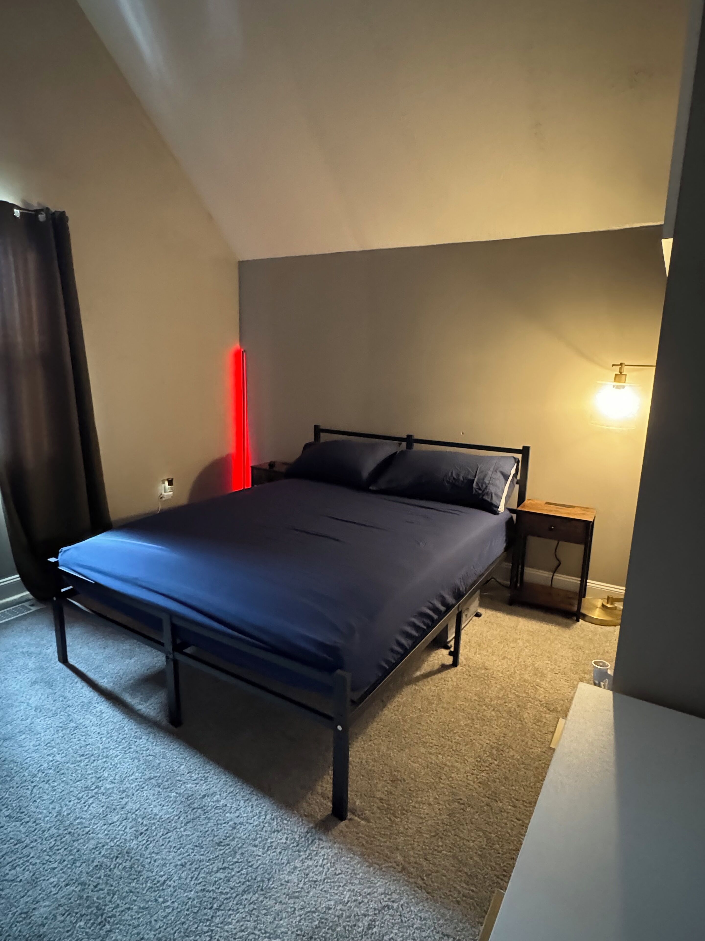 A single bed with a metal frame and dark bedding in a room with beige walls and a carpeted floor.