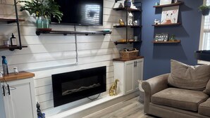 Una sala de estar con chimenea, piso de madera, un sofá, estanterías con objetos decorativos y un televisor montado en la pared.