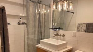 Een badkamer met een witte wastafel, houten kastje, glazen douchegordijn en een spiegel met verlichting.
