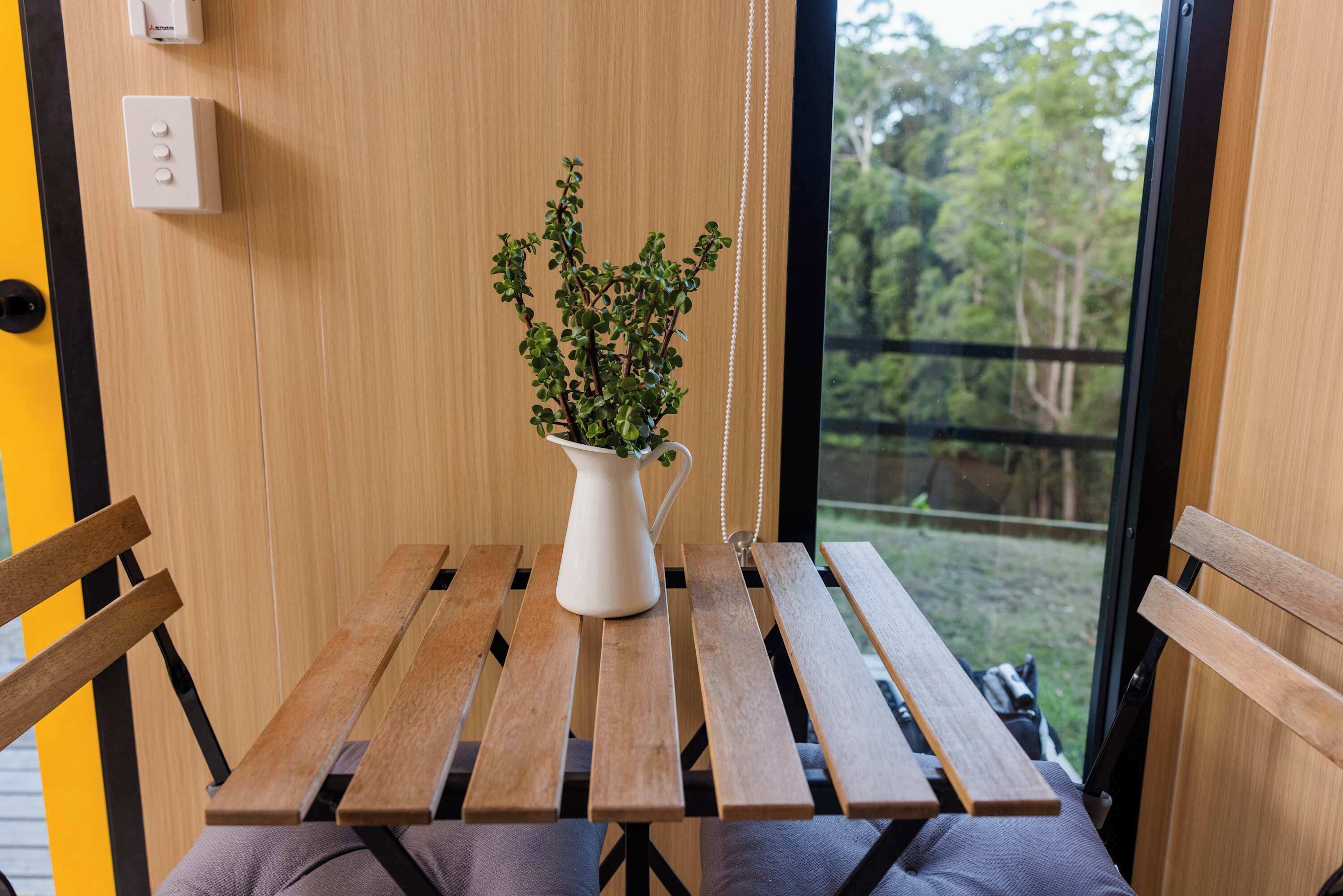 Een houten tafel met een witte kan en groene planten, een bankje, en een uitzicht op bomen door een raam.