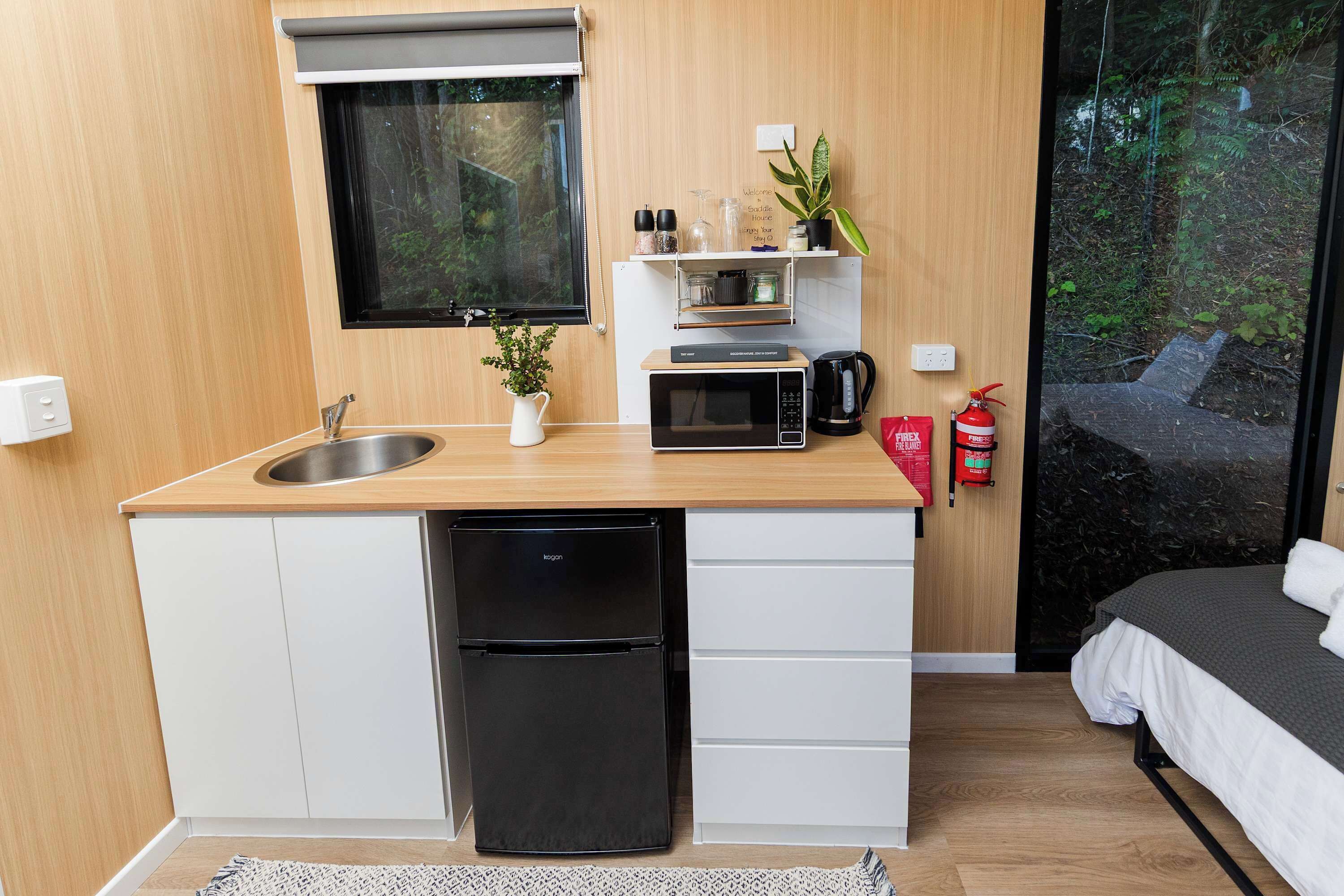 Een kleine keuken met een spoelbak, magnetron en koelkast, een kleine potplant en een brandblusser.