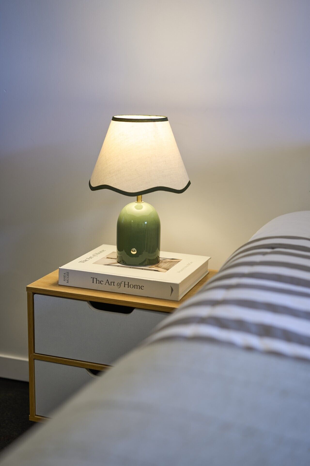 A bedside table with a lamp, a book titled 'The Art of Home,' and a striped pillow.
