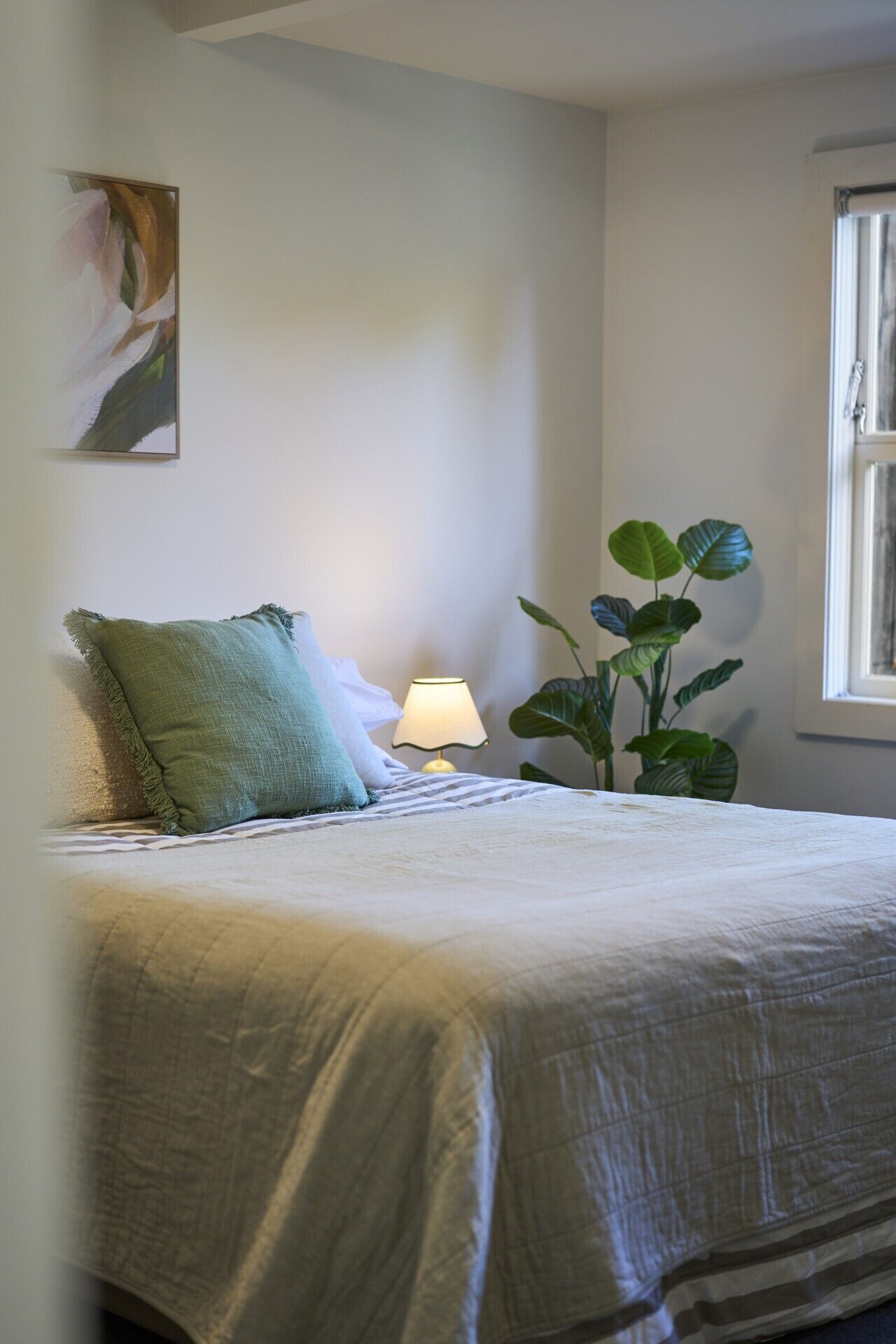 A bedroom with a bed, pillows, a bedside lamp, a plant, and a window.