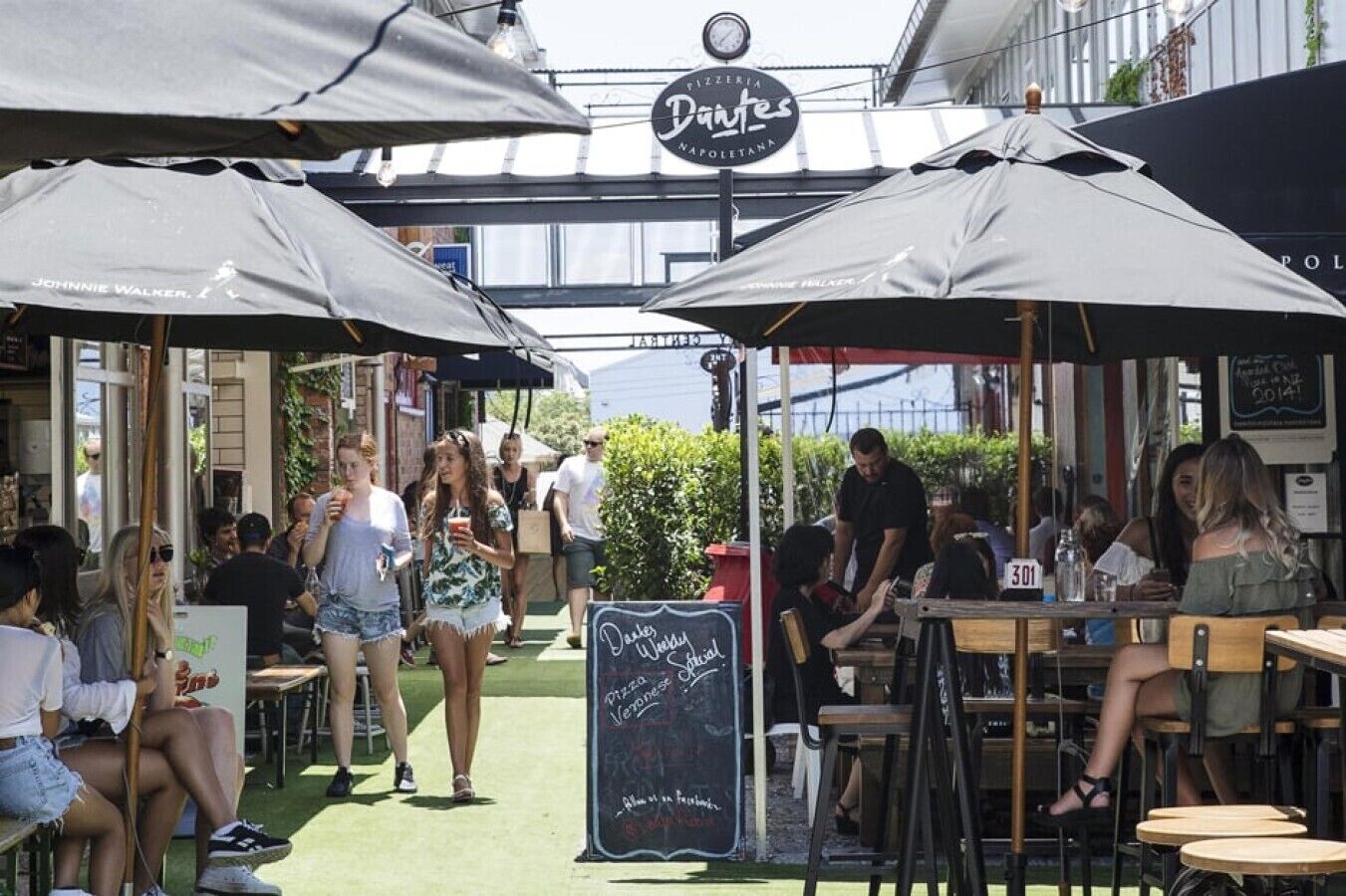 An outdoor dining area with tables and umbrellas, a chalkboard sign, and a pizzeria sign above.