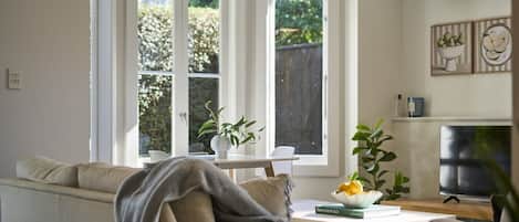 Ein Wohnzimmer mit einer Couch, einem Couchtisch mit Buch und Obst, einem Fernseher und großen Fenstern mit Blick ins Grüne.