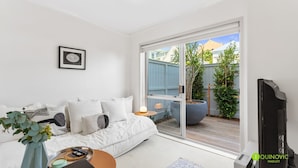 A modern living room with a white sofa, a coffee table, and a TV. There's a sliding glass door leading to a balcony with a large planter and a view of the neighborhood. - Cosy St Marys Bay Retreat with Parking (Auckland)