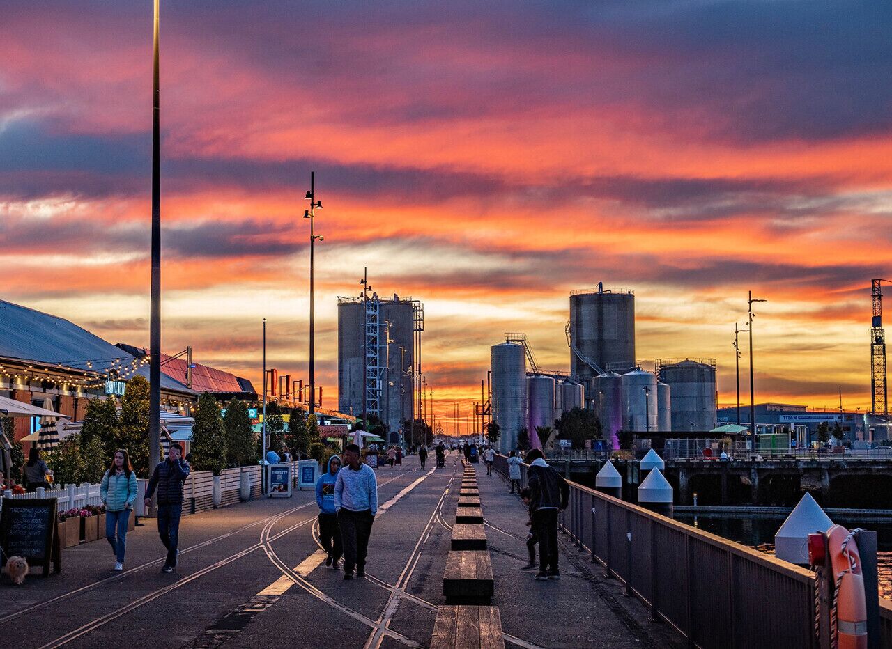A waterfront promenade with a sunset view, industrial buildings, and a pedestrian walkway.