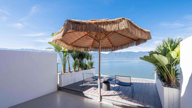 A balcony with a thatched umbrella, seating, and potted plants, overlooking a body of water and mountains. - Dali Sky & Erhai Sea View Holiday Inn (Dali)