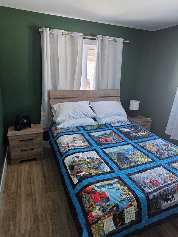 A bedroom with a wooden bed, white curtains, a bedside lamp, and a quilt with various patterns. - Cozy Beachfront Cottage Retreat w/ Loft Workspace, Stunning Views & Beach Access (King's Point)