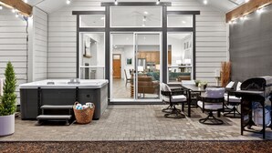 An outdoor patio with a hot tub, seating area, and a view into the house. - Downey Pines: Luxury 5bd Mountain Retreat ~ Close to Bachelor & Sunriver (Bend)
