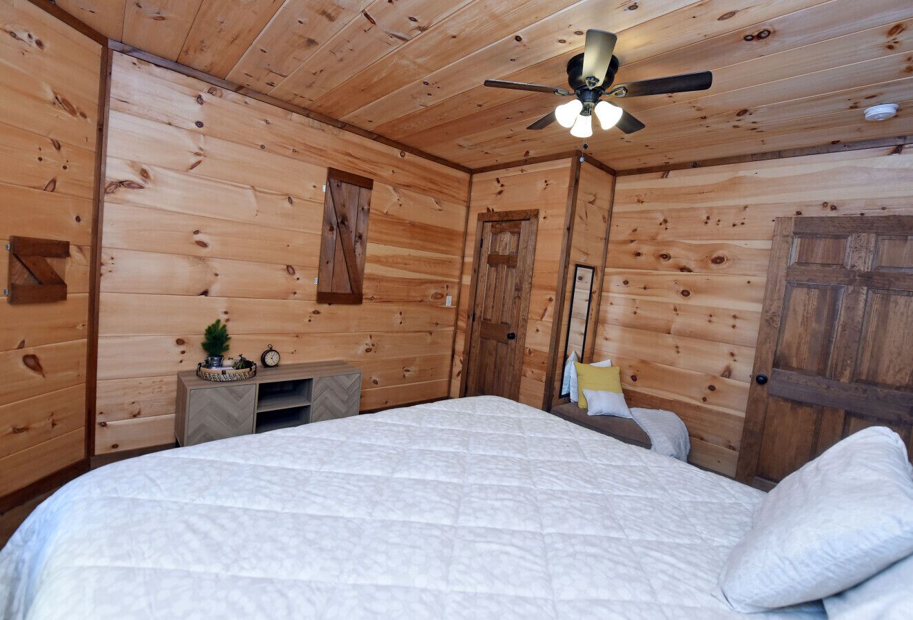 A wooden cabin bedroom with a bed, a nightstand, a dresser, a ceiling fan, and a small plant.
