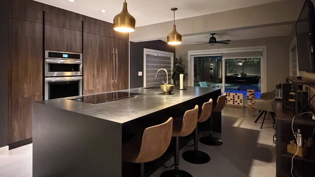 A modern kitchen with dark wood cabinets, stainless steel appliances, a central island with bar stools, pendant lighting, and a view of an outdoor pool area. - Casa Solea Miami • Pool Home w/ Workspaces + Master Office & Daybed (Miami)
