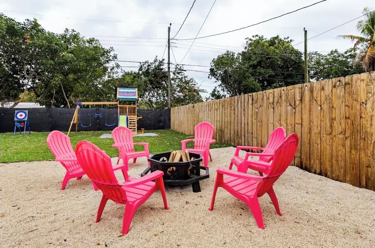 A backyard with a fire pit and pink chairs arranged around it.