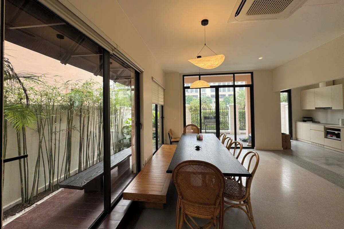 Un espace de repas moderne avec une longue table, des chaises en osier et de grandes fenêtres donnant sur un paysage verdoyant.