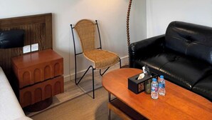 A hotel room with a wooden headboard, a wicker chair, a black leather sofa, a wooden coffee table with water bottles, a floor lamp, and a red artwork on the wall. - Calmstay-Soluna Pearl Villa-Ocean Park (Nghia Tru)